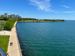 A photo of Lake Michigan in Milwaukee. The sky is bright blue with wispy clouds. The lake water is blue green and dominates the right side of the picture, but there is a small peninsula jutting out into it, covered with grass and trees. On the left side of the photo, a walking path runs along the lakefront. Several people are walking and running on the path. In the distance on the left side of the photo, you can see the city skyline.