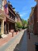 Looking down Elfreth's Alley, a historic street in Philadelphia, Pennsylvania. The sky is bright blue with wispy clouds. The center of the street is cobblestones, and to either side of the stones, the street is brick. The cobbled section is lined with red bollards. The street is narrow and lined with early American homes on either side. The homes are all red brick, some with off-white (yellow-ish) wooden trim. The first home on the left has a green door and a bicentennial flag on a pole.